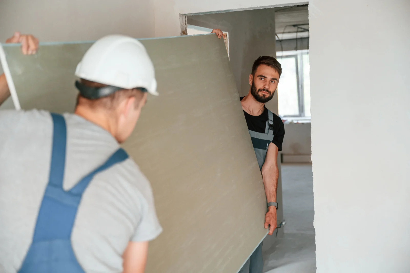 Zwei Trockenbauer tragen eine Gipskartonplatte in einen Raum im Innenausbau